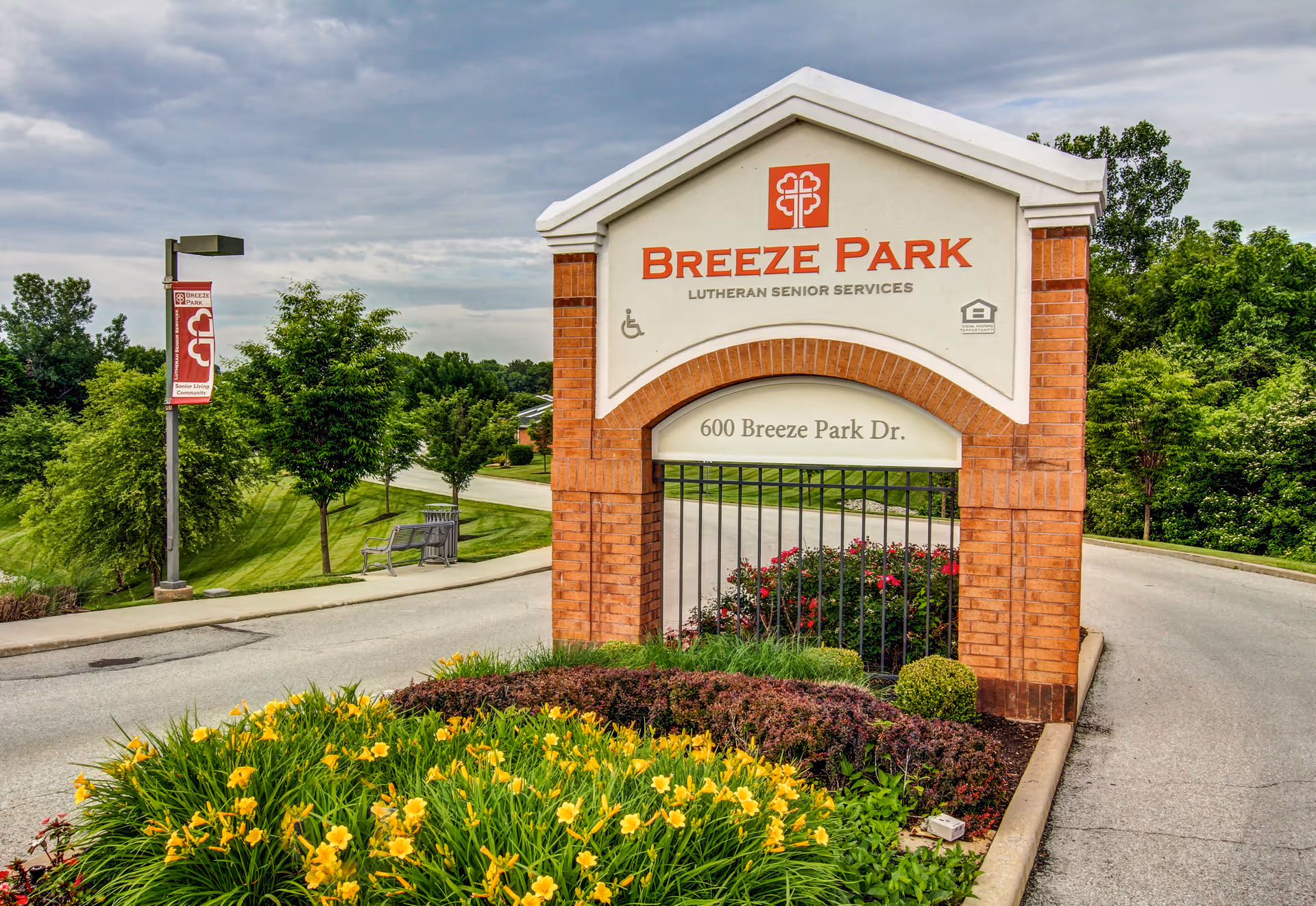 Entrance sign for Breeze Park Lutheran Senior Services located at 600 Breeze Park Dr., surrounded by landscaped flowers and greenery with a road curving around the sign under a cloudy sky.