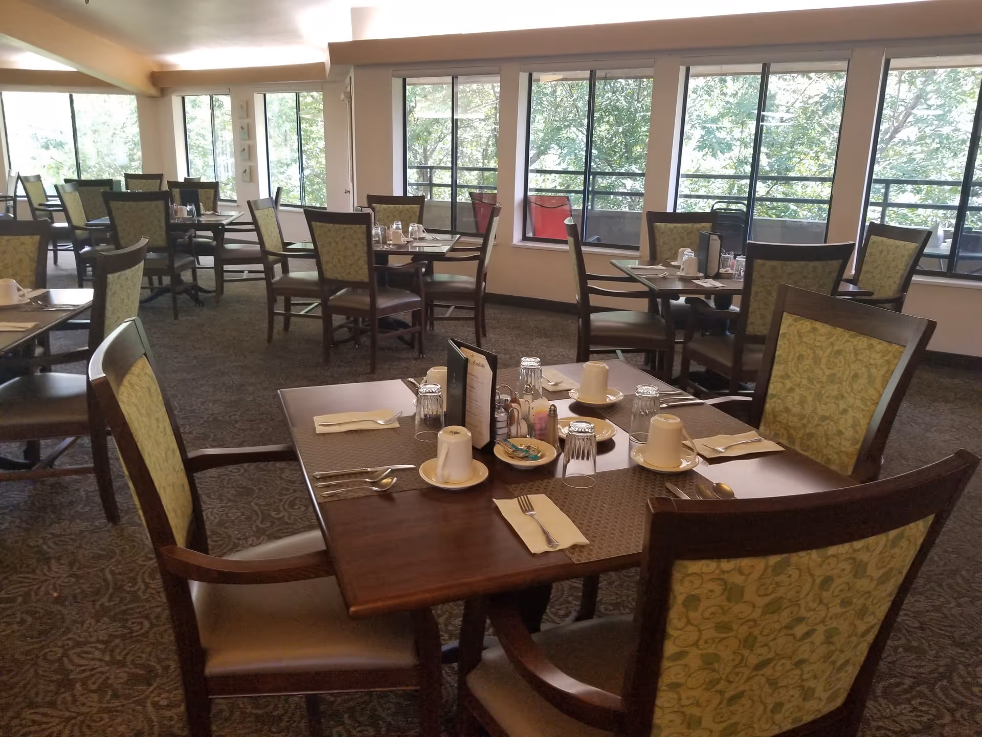 A dining room with multiple wooden tables and chairs arranged neatly. Each table is set with cups, plates, utensils, napkins, and condiments. Large windows along one wall allow natural light to fill the room and offer a view of green trees outside.