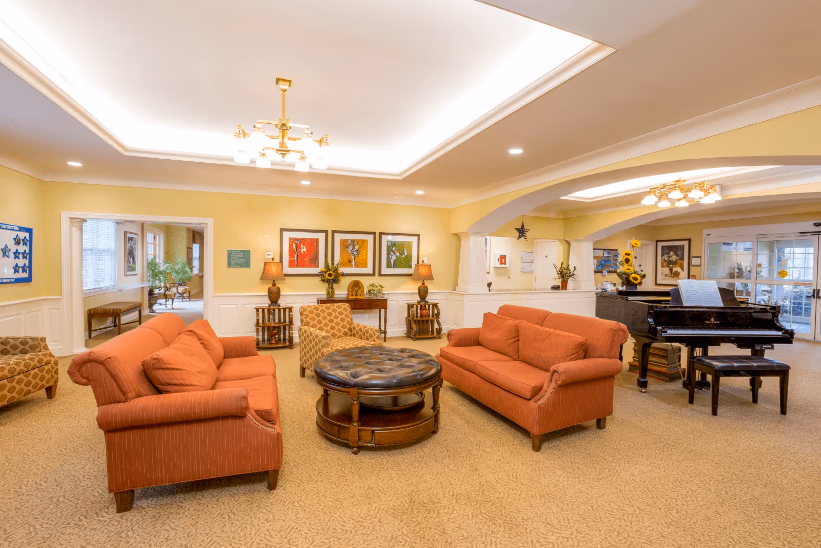 A spacious and warmly lit senior living facility common area with two orange sofas, a patterned armchair, a round tufted coffee table, and a black grand piano. The walls are painted yellow with white wainscoting, decorated with framed artwork and table lamps. There is a chandelier on the ceiling and large windows letting in natural light.