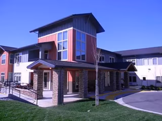 Exterior view of a two-story assisted living facility building with a covered entrance supported by stone pillars, large windows, and a well-maintained lawn under a clear blue sky.