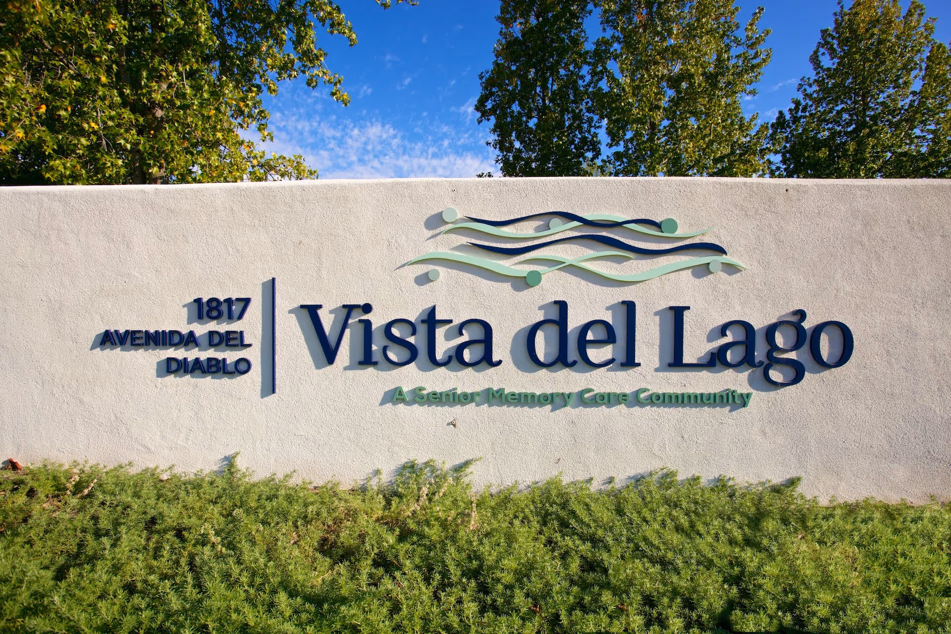 Outdoor sign for Vista del Lago, a senior memory care community, mounted on a white wall with green shrubs in front and trees in the background under a blue sky.