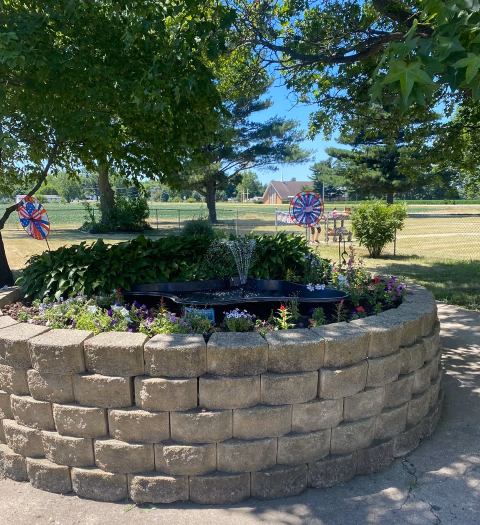 A circular raised stone planter containing a small fountain and colorful flowers beneath trees with a grassy field and chain-link fence in the background.
