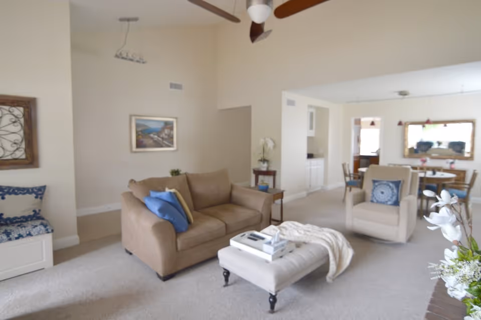 A bright and spacious living room with beige walls and carpet. The room features a tan sofa with blue and beige pillows, a beige armchair with a blue patterned pillow, and a tufted ottoman with a tray and a blanket. There is a ceiling fan with wooden blades, a framed painting on the wall, and a dining area with a round table and chairs in the background. Decorative elements include a floral arrangement and a small side table with a plant.