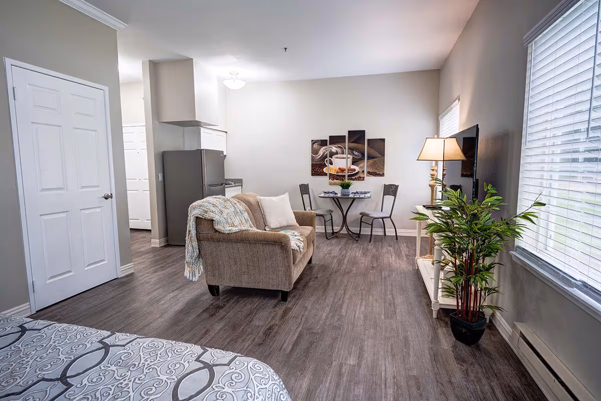 Interior view of a senior living facility room at Gig Harbor Court featuring a small living area with a beige loveseat draped with a blanket, a round table with two chairs against the wall, a flat-screen TV on a white stand with a lamp and a potted plant beside it, and a bed partially visible in the foreground. The room has wood flooring, light gray walls, and windows with white blinds.