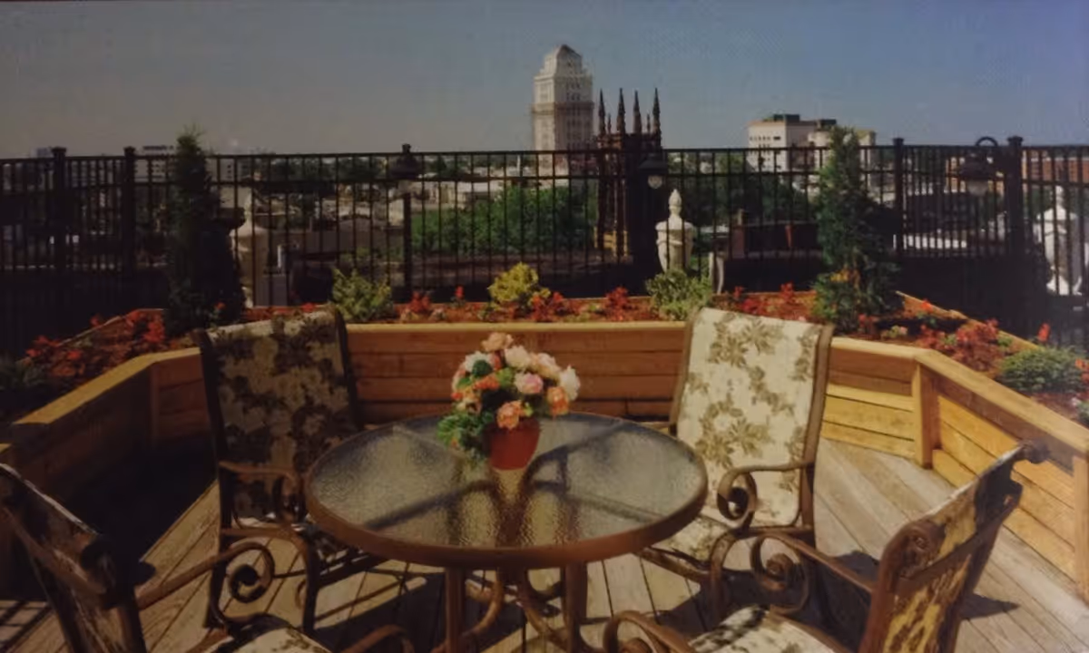 Outdoor patio area with a round glass table and four cushioned chairs with floral patterns. A flower arrangement in a red pot sits on the table. The patio is surrounded by wooden planters with various plants and flowers, and a black metal fence encloses the area. In the background, a cityscape with buildings and a tall tower is visible under a clear sky.