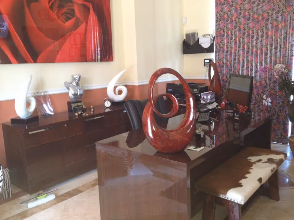 An office or reception area with a dark wooden desk and matching cabinet. The desk has a computer monitor, office supplies, and a large red abstract sculpture. The cabinet behind the desk holds decorative items including white curved sculptures, a silver bust, and a small clock. A large red rose painting hangs on the wall above the cabinet, and floral patterned curtains cover the window on the right side. A bench with a cowhide cushion is positioned in front of the desk.
