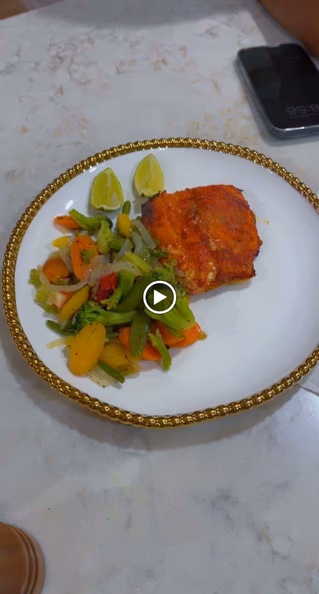 A white plate with a gold decorative rim containing a serving of cooked mixed vegetables and a piece of grilled or baked fish, accompanied by two lime wedges. The plate is on a light-colored marble countertop with a smartphone and part of a person's arm visible in the background.