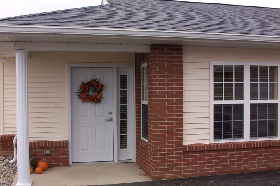 Entrance to a residential unit with a white door decorated with a fall-themed wreath, beige siding, red brick accents, a white column, and three small pumpkins on the ground near the corner.