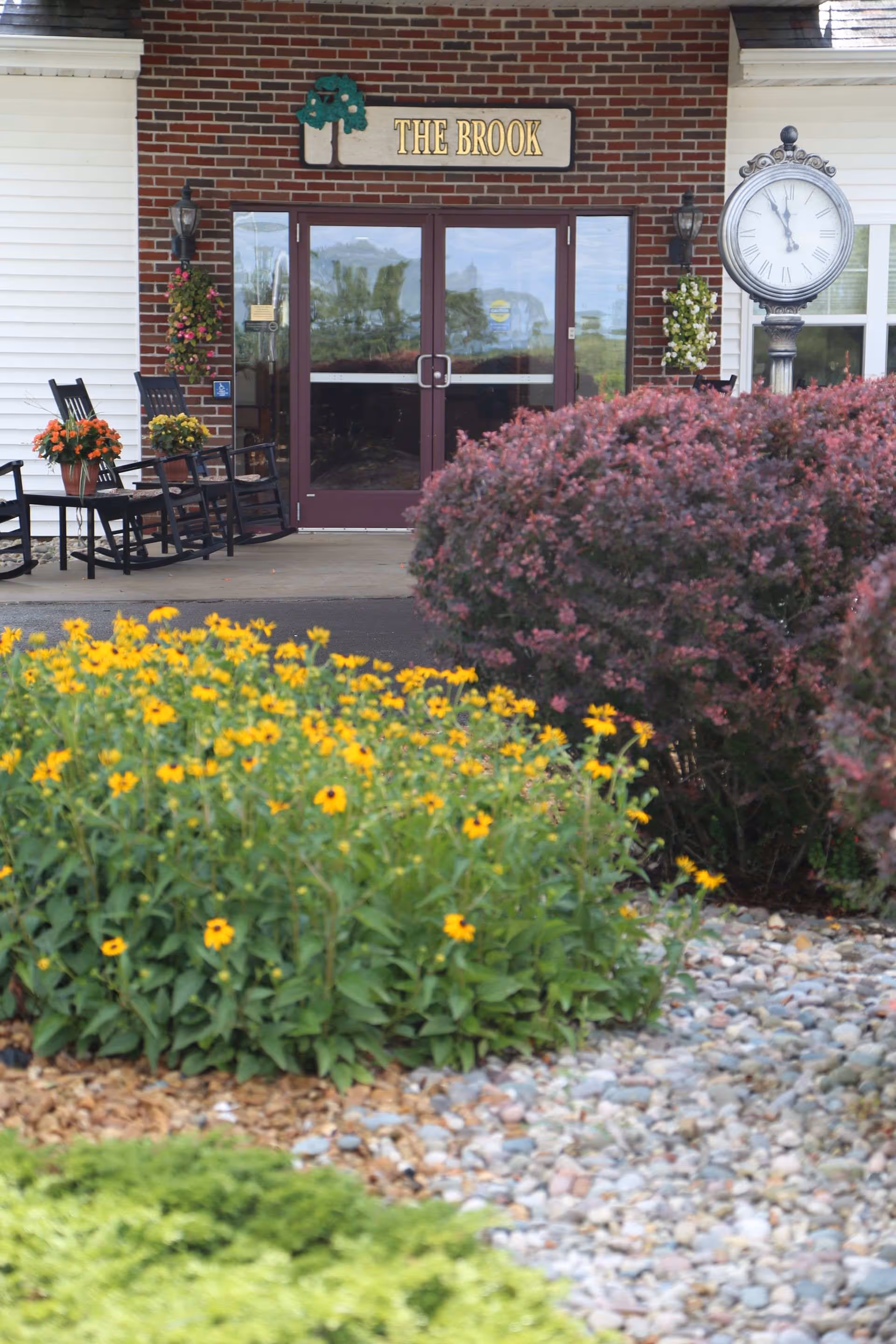 Front entrance of The Brook senior living facility with double glass doors, rocking chairs, a large outdoor clock, and landscaped flower beds.