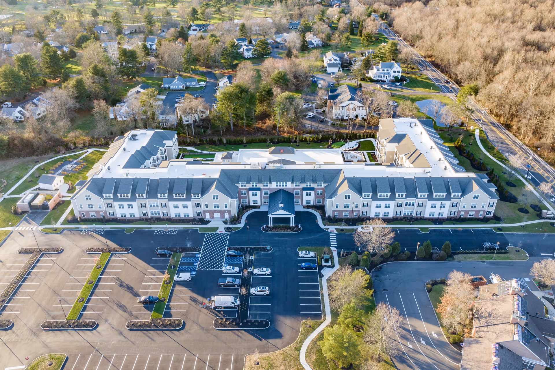 Aerial view of a large senior living facility building with a front entrance, parking lot, and surrounding trees and homes.