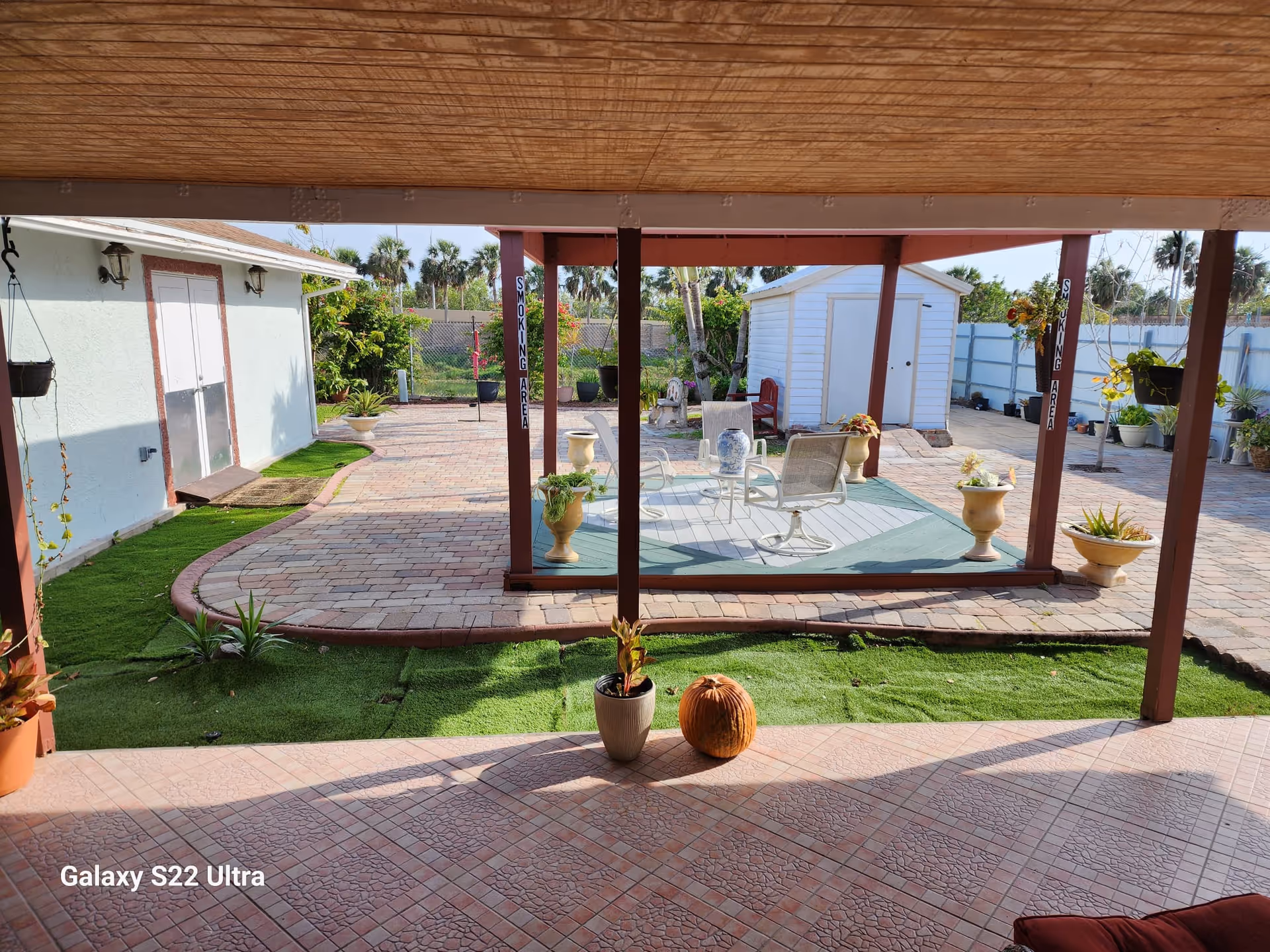 Outdoor patio area with tiled flooring and artificial grass patches, featuring several potted plants and a pumpkin. There is a covered seating area with chairs and a small table on a raised platform. In the background, there is a white shed and a fenced garden area with palm trees and other greenery. Two vertical signs on wooden posts indicate a smoking area.