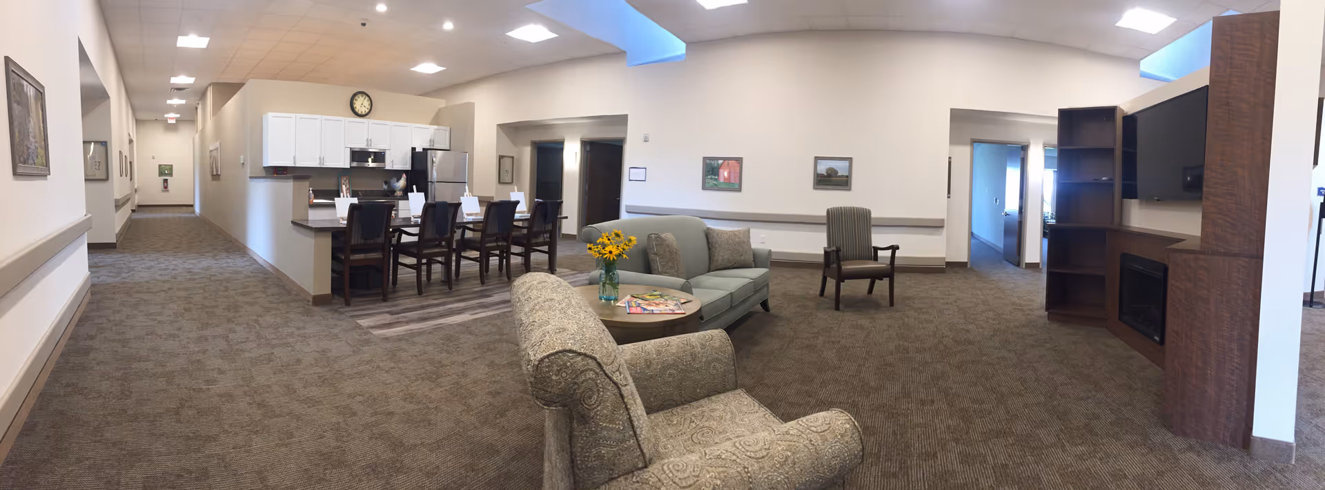 Interior view of a senior living facility common area featuring a cozy seating arrangement with two armchairs, a loveseat, a round table with a vase of yellow flowers and magazines, a large flat-screen TV mounted above a wooden fireplace, and a kitchen area with a counter and four high chairs. The space has carpeted floors, white walls with framed pictures, and a hallway leading to other rooms.