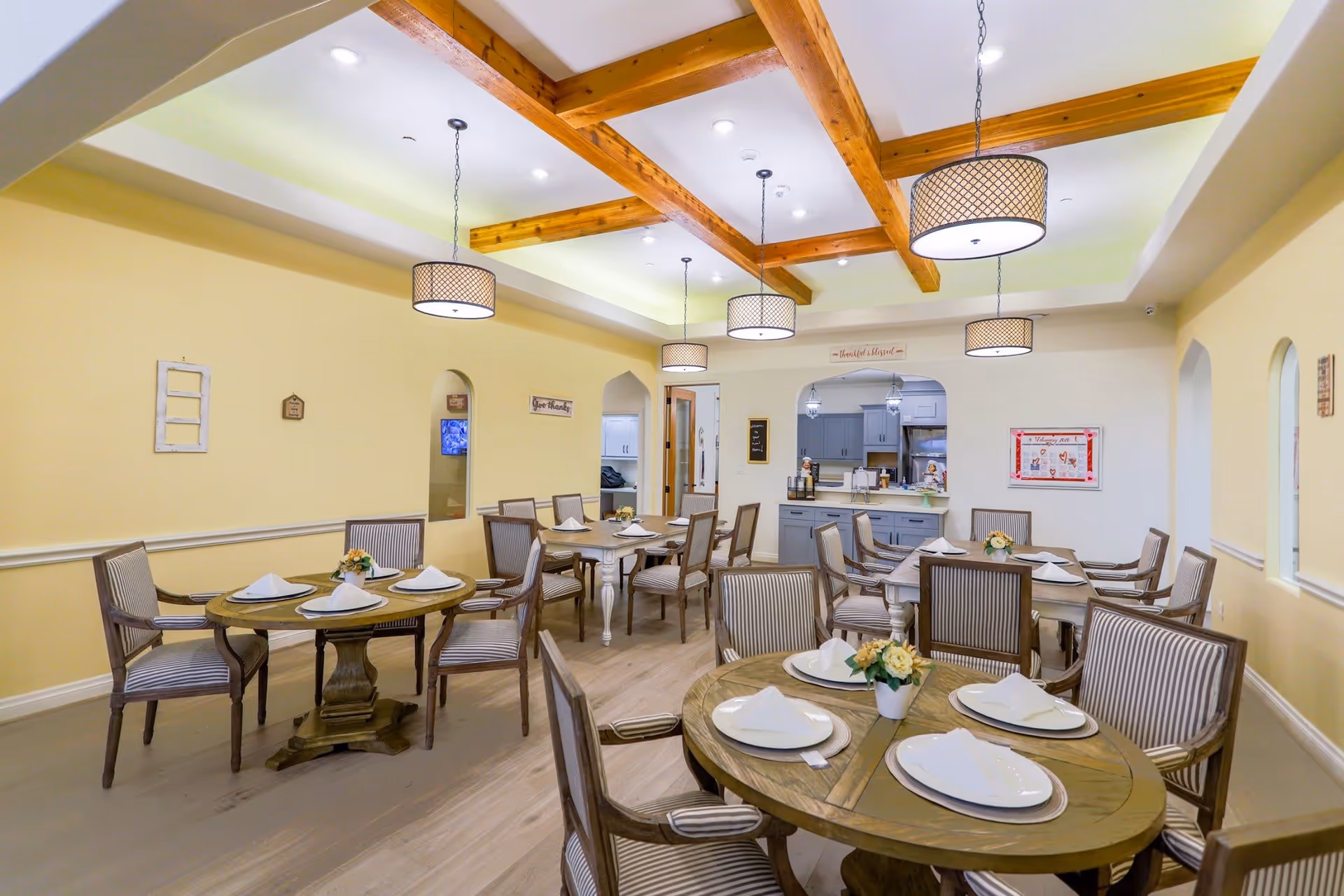 Bright dining room with round tables set with plates and napkins, striped chairs, pendant lights, and exposed wooden ceiling beams.