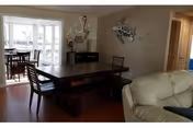 Interior view of a senior living facility showing a dining area with a dark wooden table and chairs, a beige leather sofa in the foreground, and a bright sunroom with additional seating in the background. The walls are decorated with a metal fish sculpture and the word 'Joy'.