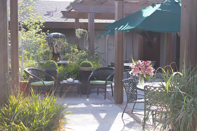 Outdoor patio area with wicker chairs and a small table set with a teapot and cups. There is a green umbrella shading a round metal table with a vase of pink lilies. The area is surrounded by plants and greenery, with a decorative water fountain in the background.