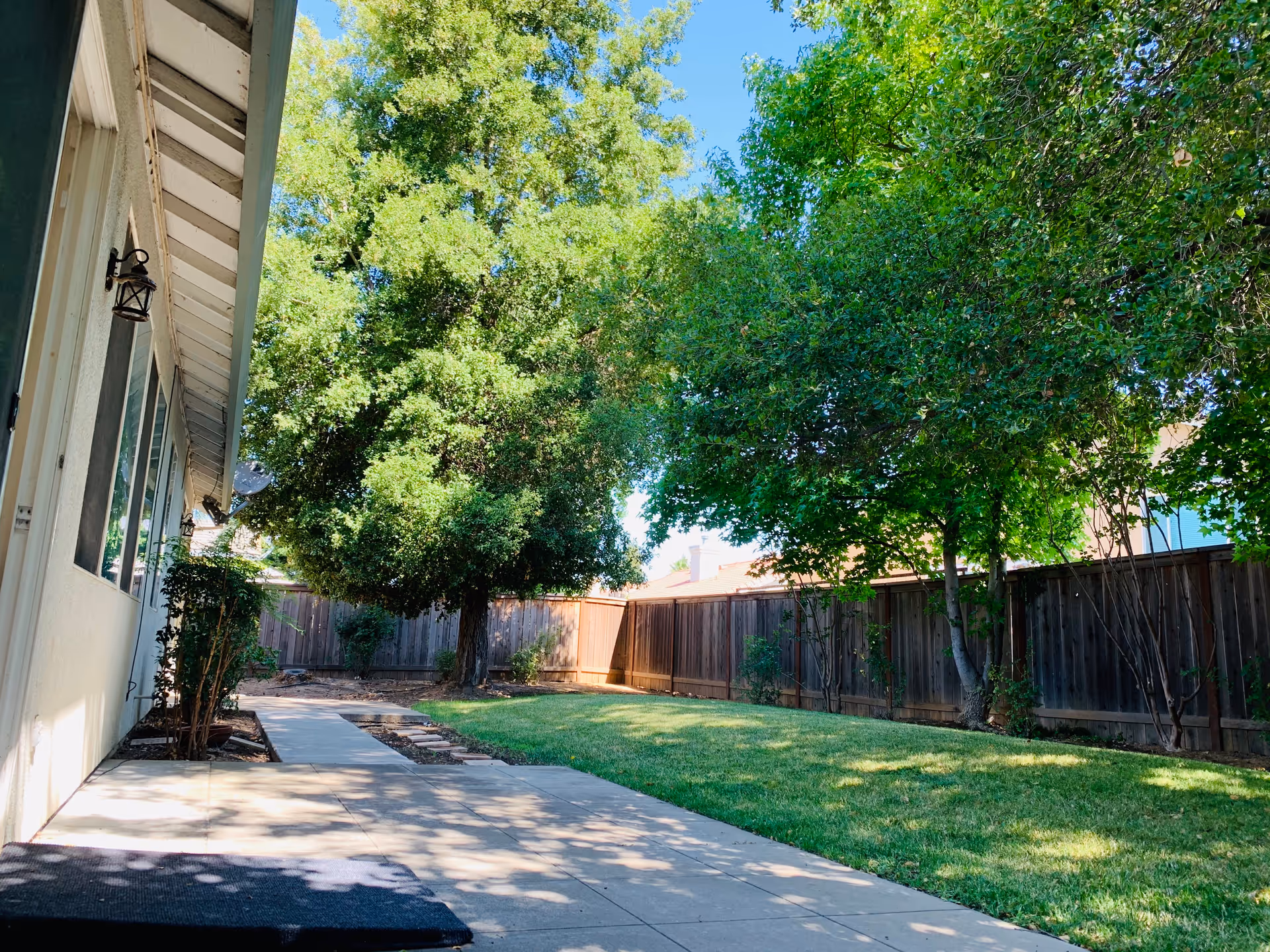 Outdoor view of a fenced backyard area with a concrete pathway, green grass, and several large leafy trees providing shade. The side of a building with windows and wall-mounted lanterns is visible on the left side.