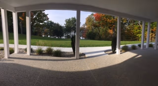 Covered outdoor patio area with white pillars overlooking a green lawn and trees with autumn foliage in the background under a clear sky.