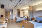 A clean and simple nursing facility room with two hospital beds, each equipped with side rails and a small overbed table. The room has a light-colored floor and walls, a ceiling-mounted curtain divider between the beds, a small television mounted on the wall, and a few chairs along the wall near the entrance.
