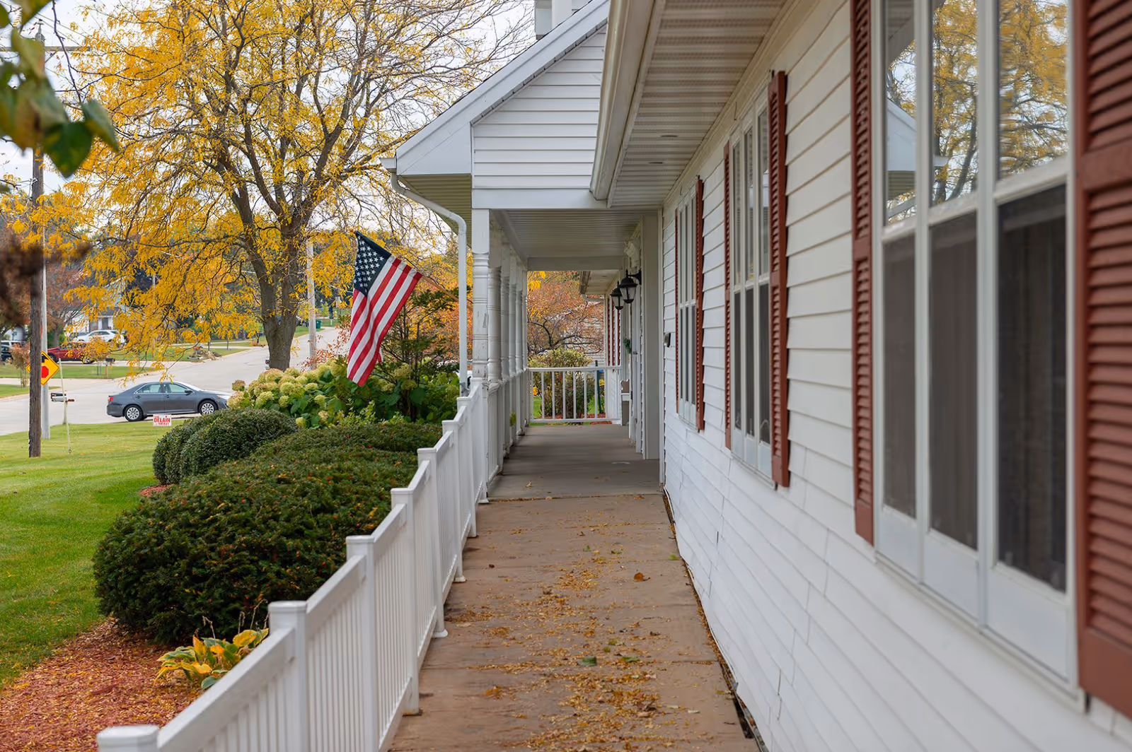 Covered porch walkway of a white building with an American flag, white railing, and autumn trees in the background.