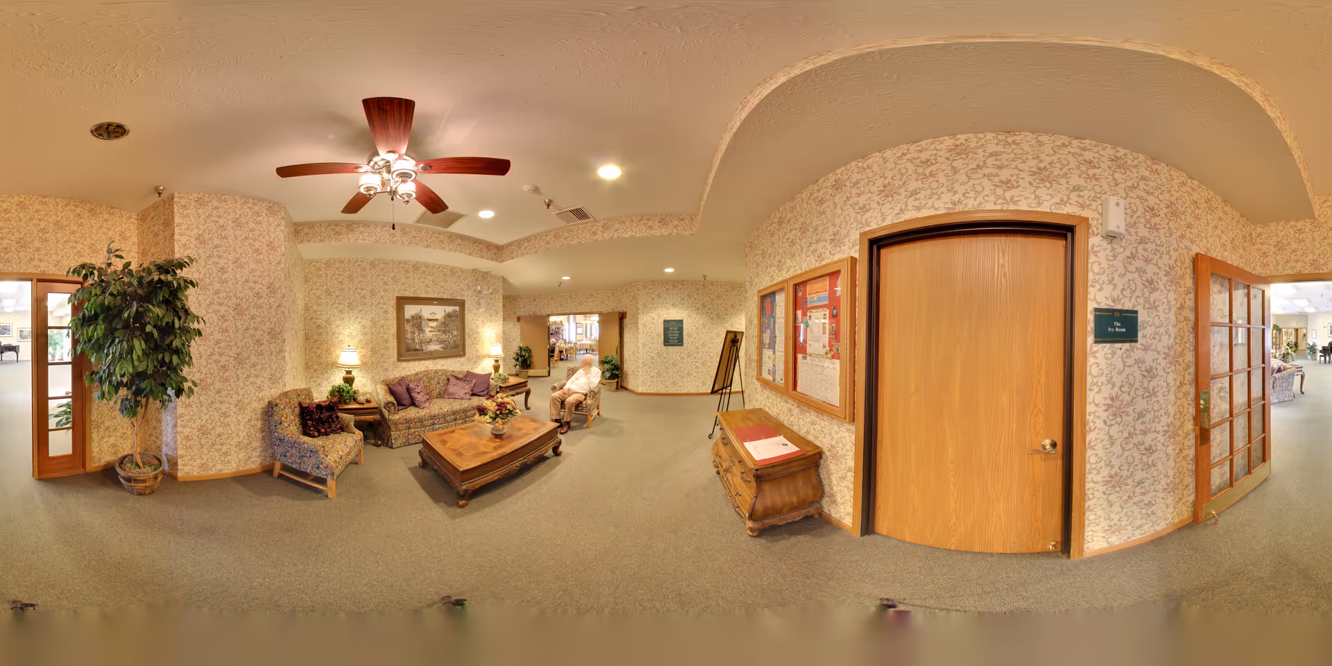A cozy common area in a senior living facility with floral wallpaper, a ceiling fan with lights, a wooden coffee table, a sofa with purple cushions, an armchair, a potted plant, and a bulletin board on the wall. There is a wooden door and glass-paneled doors leading to other rooms, with a person sitting on a chair in the background.
