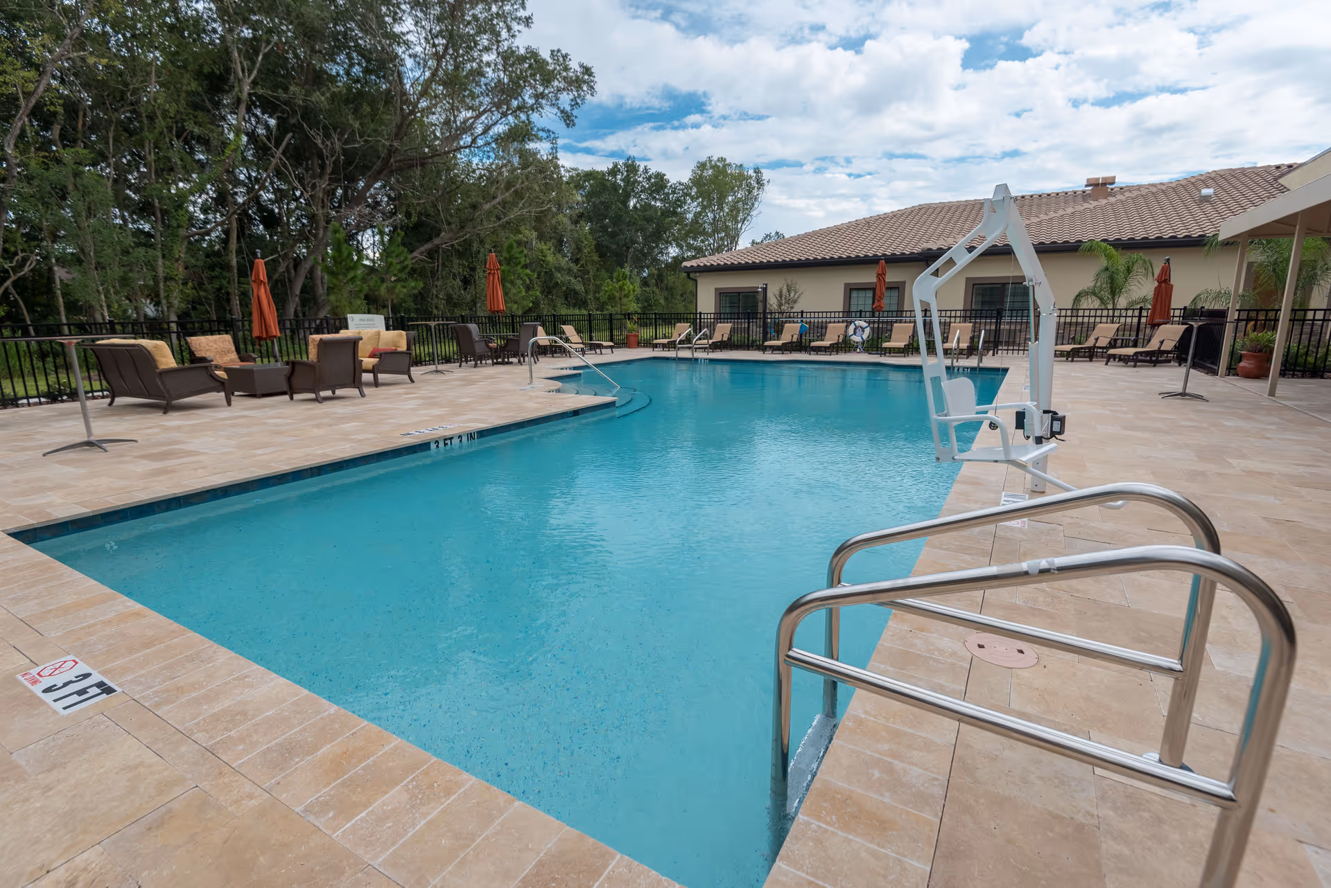 Outdoor swimming pool with tiled deck, metal handrails, a pool lift, lounge chairs and umbrellas beside a low building and trees.