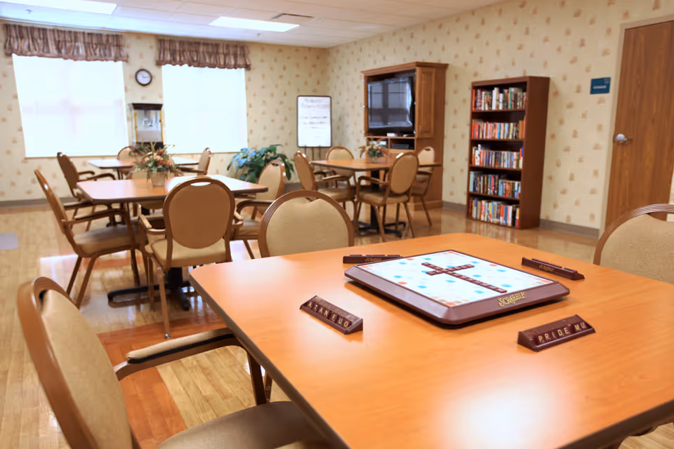 A senior living community activity room with tables and chairs, a Scrabble board on the nearest table, bookshelves and a TV in the background.