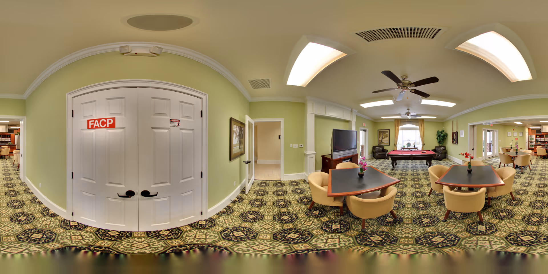A spacious common area in a senior living facility with green walls and patterned carpet. The room features two tables with beige chairs and small flower vases, a large flat-screen TV on a wooden stand, and a pool table near a window with curtains. Ceiling fans and recessed lighting are visible on the ceiling. Double white doors with a red FACP sign are on one side, and there are additional rooms visible through open doorways.