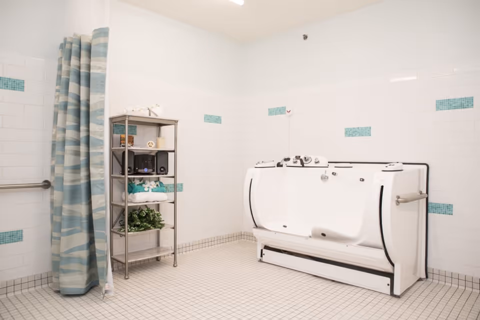 A clean, tiled bathroom with a walk-in bathtub on the right side, a metal shelving unit with towels, a radio, and decorative items in the center, and a blue and white patterned shower curtain on the left side.
