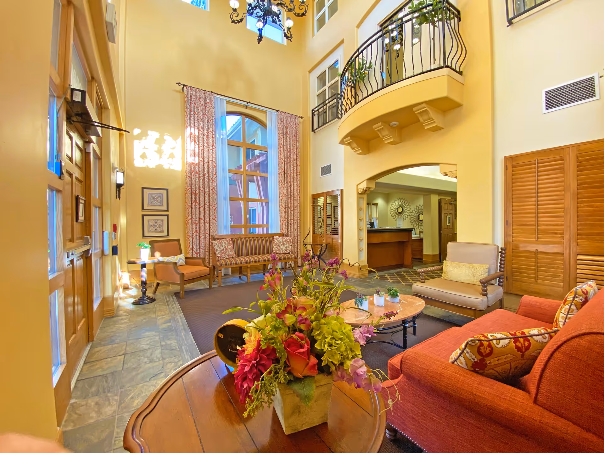 A bright and spacious living room area with high ceilings and a balcony above. The room features a red sofa with patterned pillows, a beige armchair, a wooden table with a colorful flower arrangement, and additional seating near a large window with patterned curtains. The walls are painted a warm yellow, and there is a chandelier hanging from the ceiling. The room has a welcoming and cozy atmosphere.