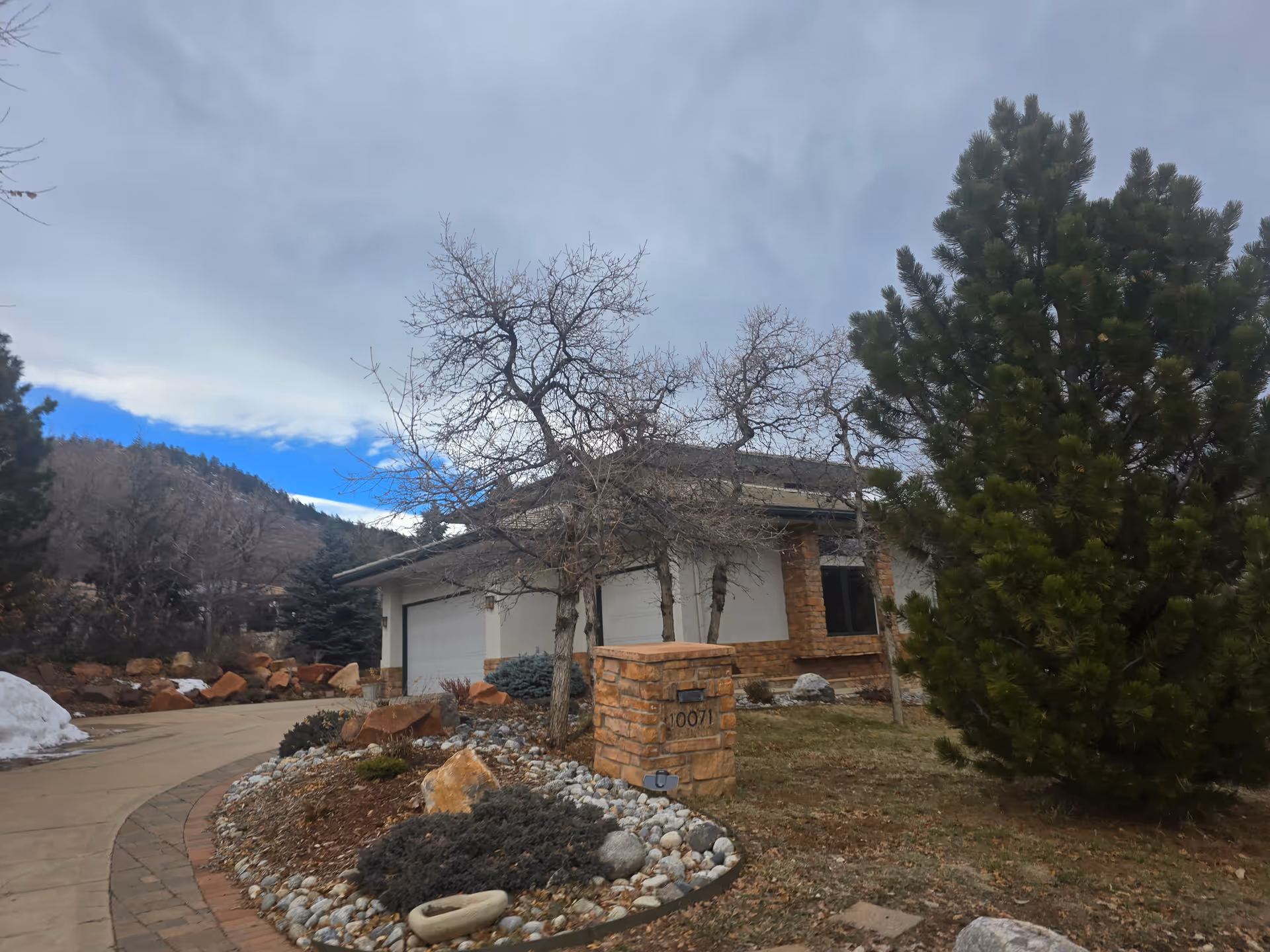 Exterior view of a single-story building with a stone and stucco facade, a garage door, and a driveway curving to the left. There are leafless trees and evergreen shrubs in the landscaped front yard, with a stone mailbox displaying the number 10071. The sky is partly cloudy with patches of blue.