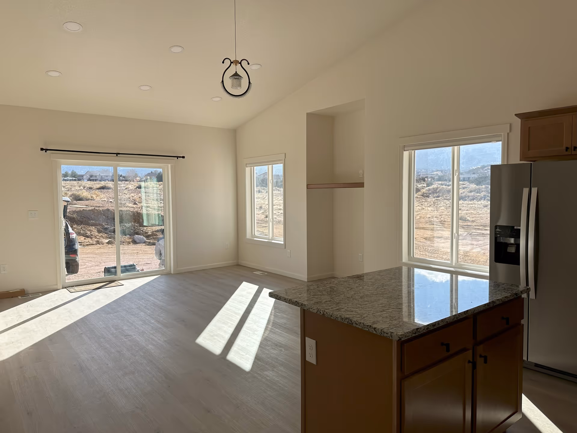 A bright, empty interior space with light-colored walls and flooring. There is a kitchen island with a granite countertop and wooden cabinets on the right side, next to a stainless steel refrigerator. The room has large windows and a sliding glass door that lets in natural light and offers a view of a dry, rocky outdoor landscape. A small pendant light hangs from the ceiling.