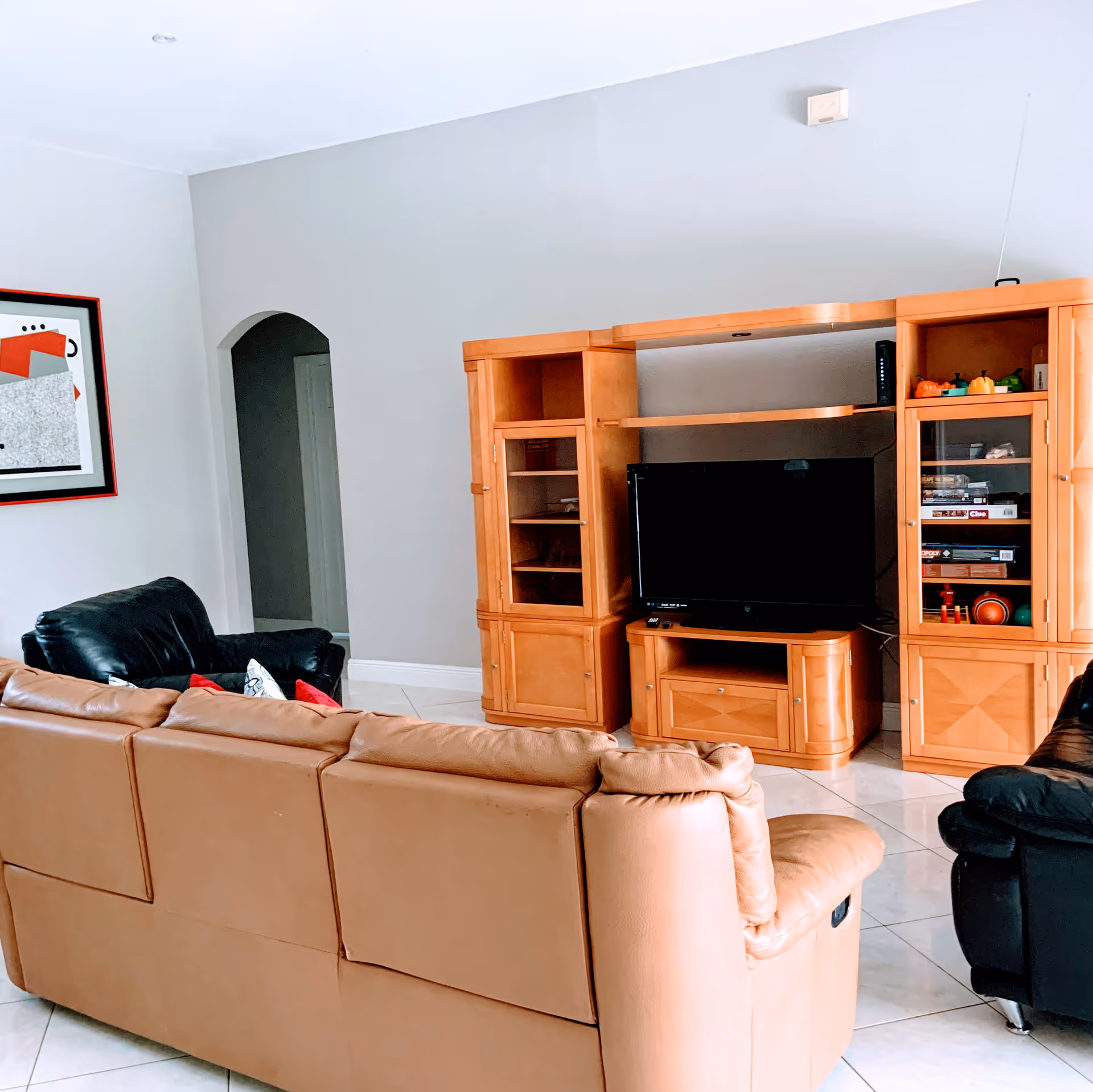 A living room with a tan leather sofa facing a wooden entertainment center that holds a flat-screen TV. There are two black leather armchairs on either side of the sofa. The entertainment center has glass cabinets with various items inside. The walls are light gray, and there is a framed abstract artwork on the left wall. The floor is tiled in white.