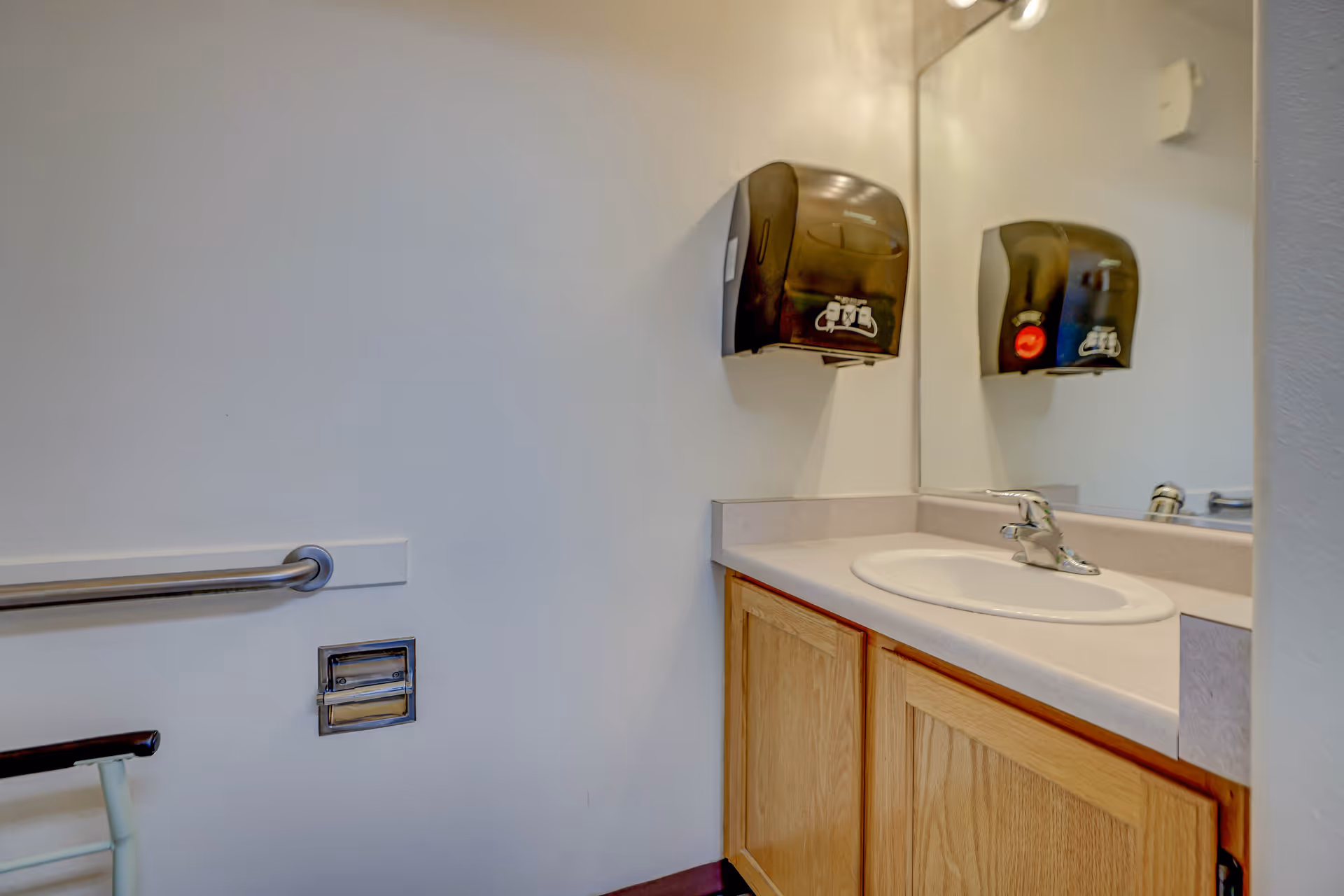 Accessible bathroom sink and wooden vanity with mirror, wall-mounted paper towel dispensers and a grab bar.