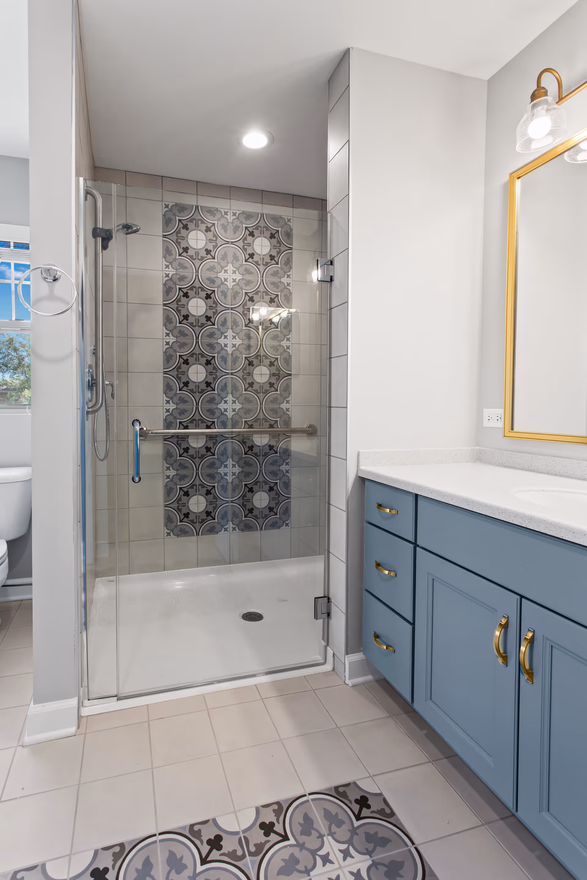 Modern bathroom with a glass-enclosed shower featuring decorative patterned tiles on the back wall. The bathroom has a blue vanity with gold handles, a white countertop, and a gold-framed mirror with a light fixture above it. The floor has light-colored tiles with a matching patterned tile rug in front of the vanity.