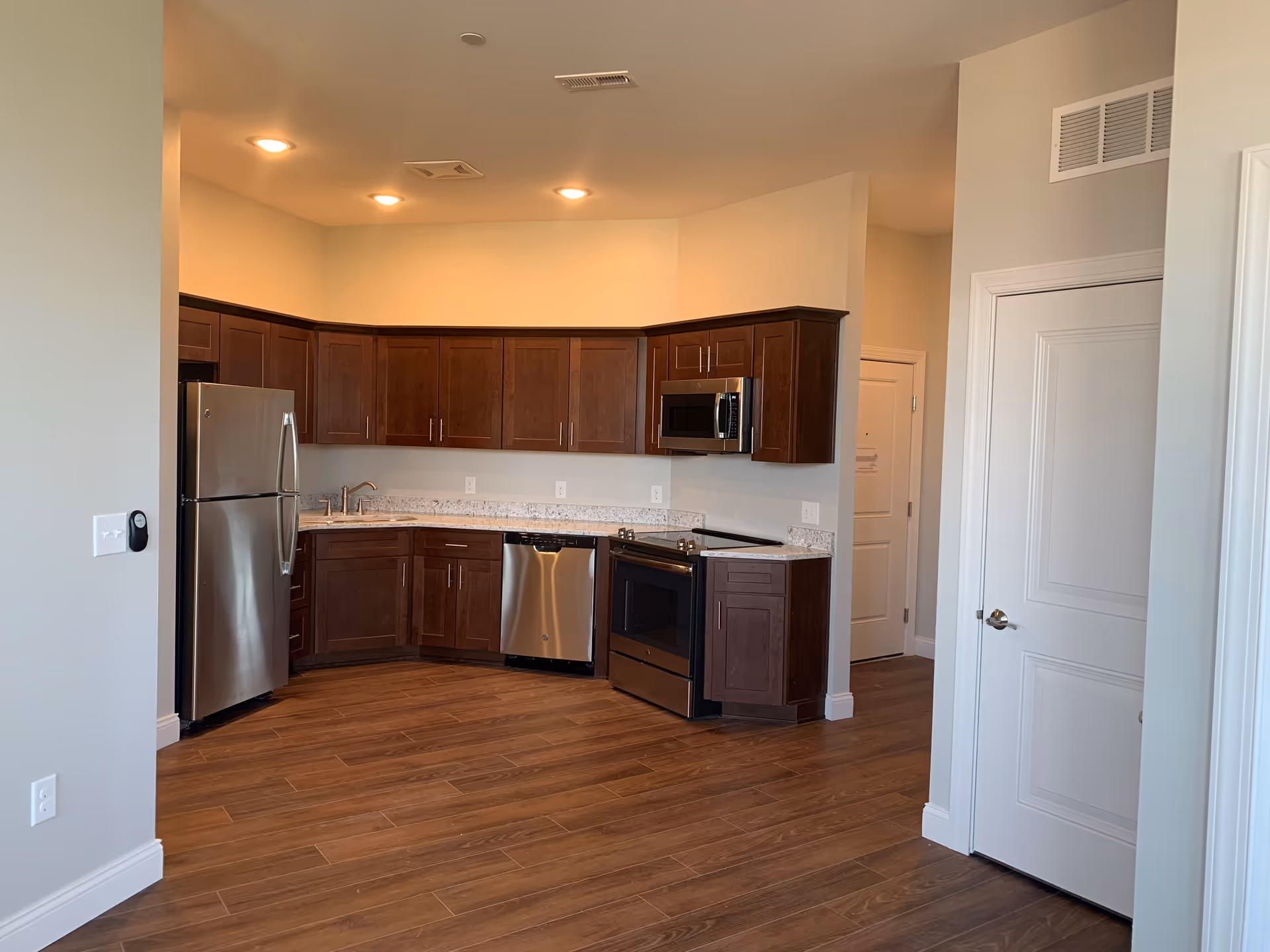 Modern kitchen with dark wood cabinets, stainless steel refrigerator, dishwasher, oven, and microwave. The kitchen has granite countertops and wood flooring, with recessed ceiling lights.
