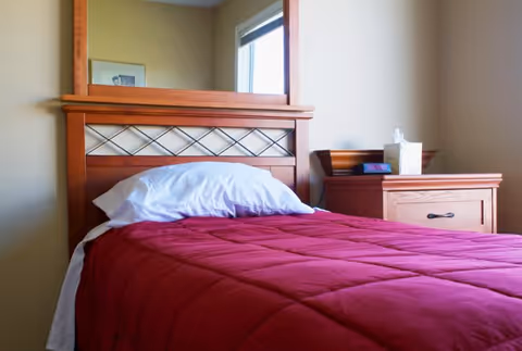 A single bed with a wooden headboard and a red quilted blanket, a white pillow, and a wooden nightstand beside it holding a tissue box and a small item, with a window letting in natural light in the background.