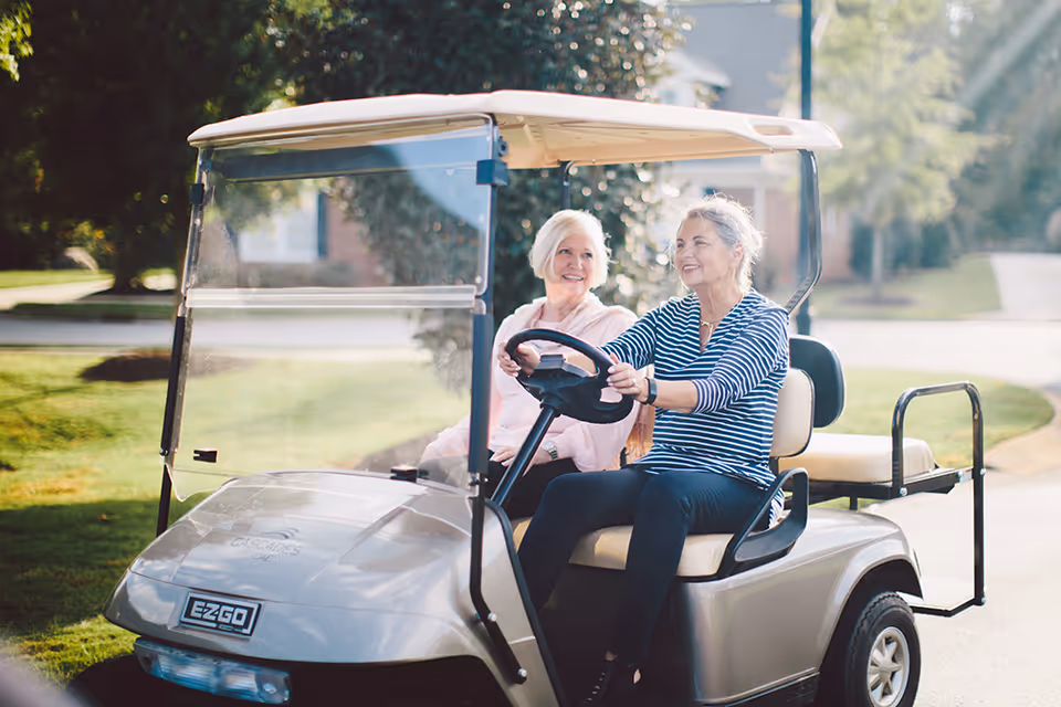 Two elderly women sitting in a golf cart outdoors on a sunny day, one driving and the other sitting beside her, both smiling and enjoying the ride.