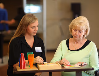 A young female caregiver and an elderly woman sitting at a table playing a board game together in an indoor setting.