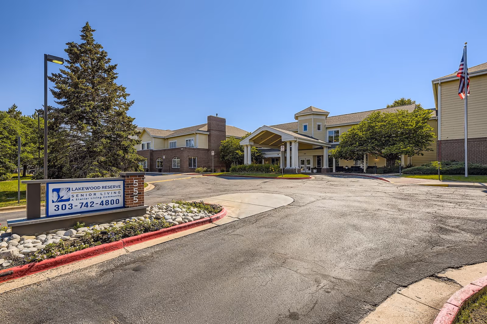 Exterior view of Lakewood Reserve Assisted Living & Memory Care facility showing the main entrance with a covered drop-off area, a large building with multiple windows, an American flag on a flagpole, and a sign with the facility's name and phone number near a tree and landscaped area.