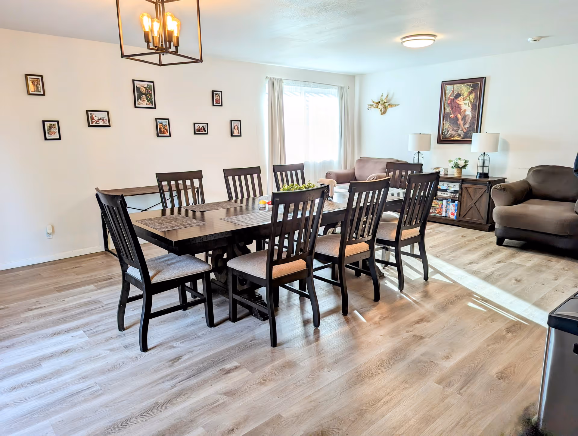 A bright dining and living area with a large wooden dining table surrounded by eight chairs. The room has light wood flooring, white walls adorned with small framed pictures, and a window with white curtains letting in natural light. In the background, there are two armchairs, a sideboard with lamps and board games, and a framed painting on the wall.
