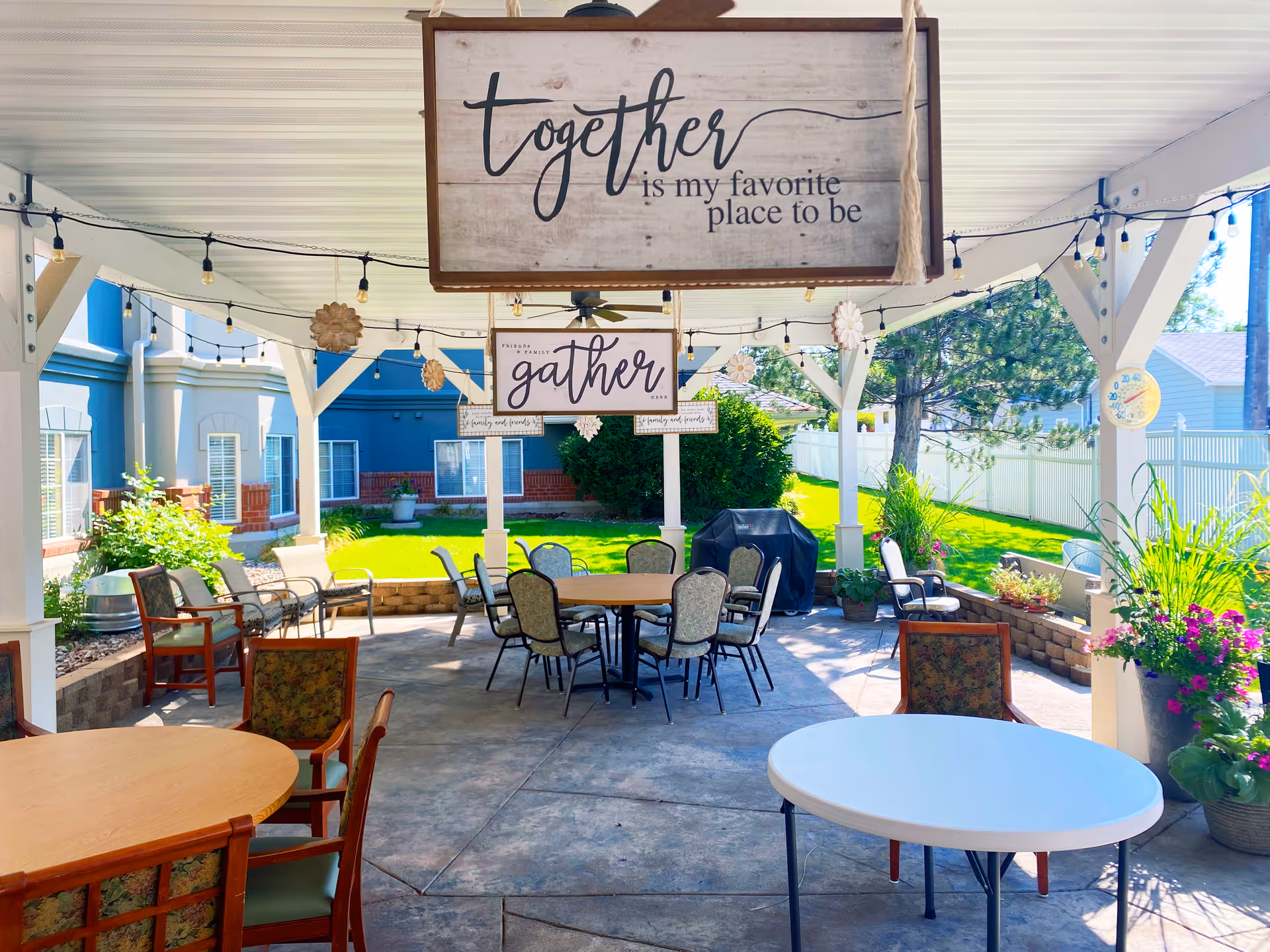 Covered outdoor patio with tables and chairs, hanging signs reading "together" and "gather," and a view of a garden and lawn.