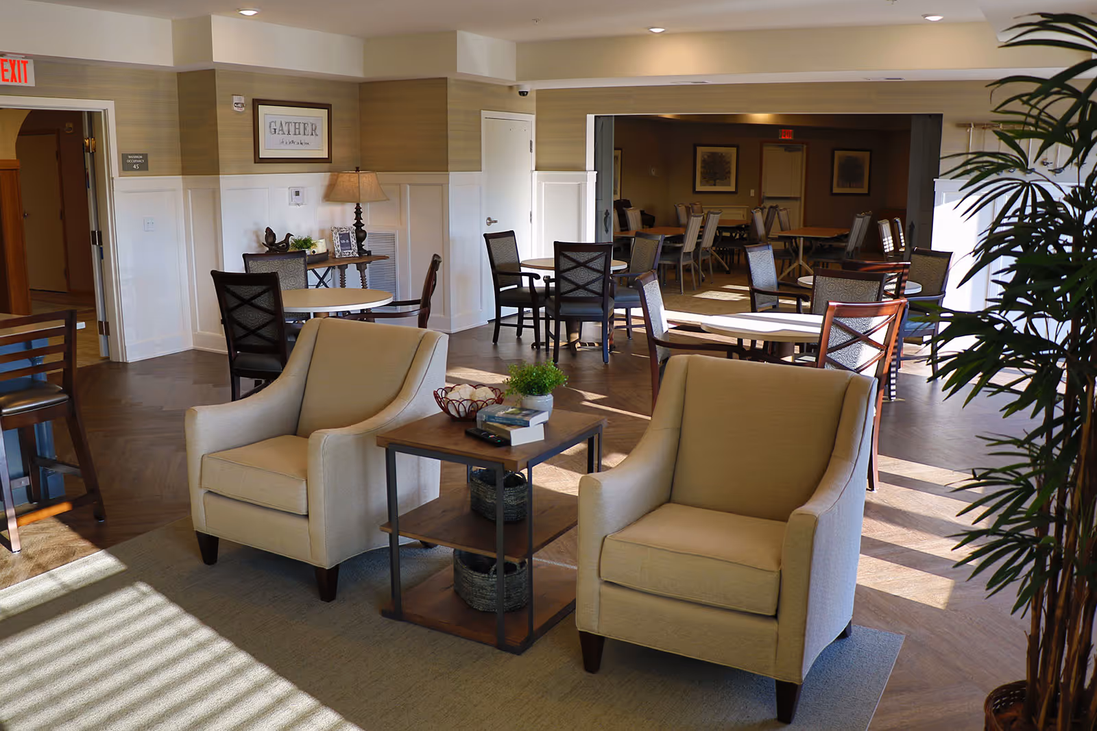 A bright and spacious common area in a senior living facility featuring two beige armchairs with a small wooden side table between them holding books and a plant. In the background, there are multiple round tables with chairs arranged for dining or socializing. The walls are decorated with framed artwork and a lamp on a small table. Sunlight streams in through windows, casting shadows on the floor.
