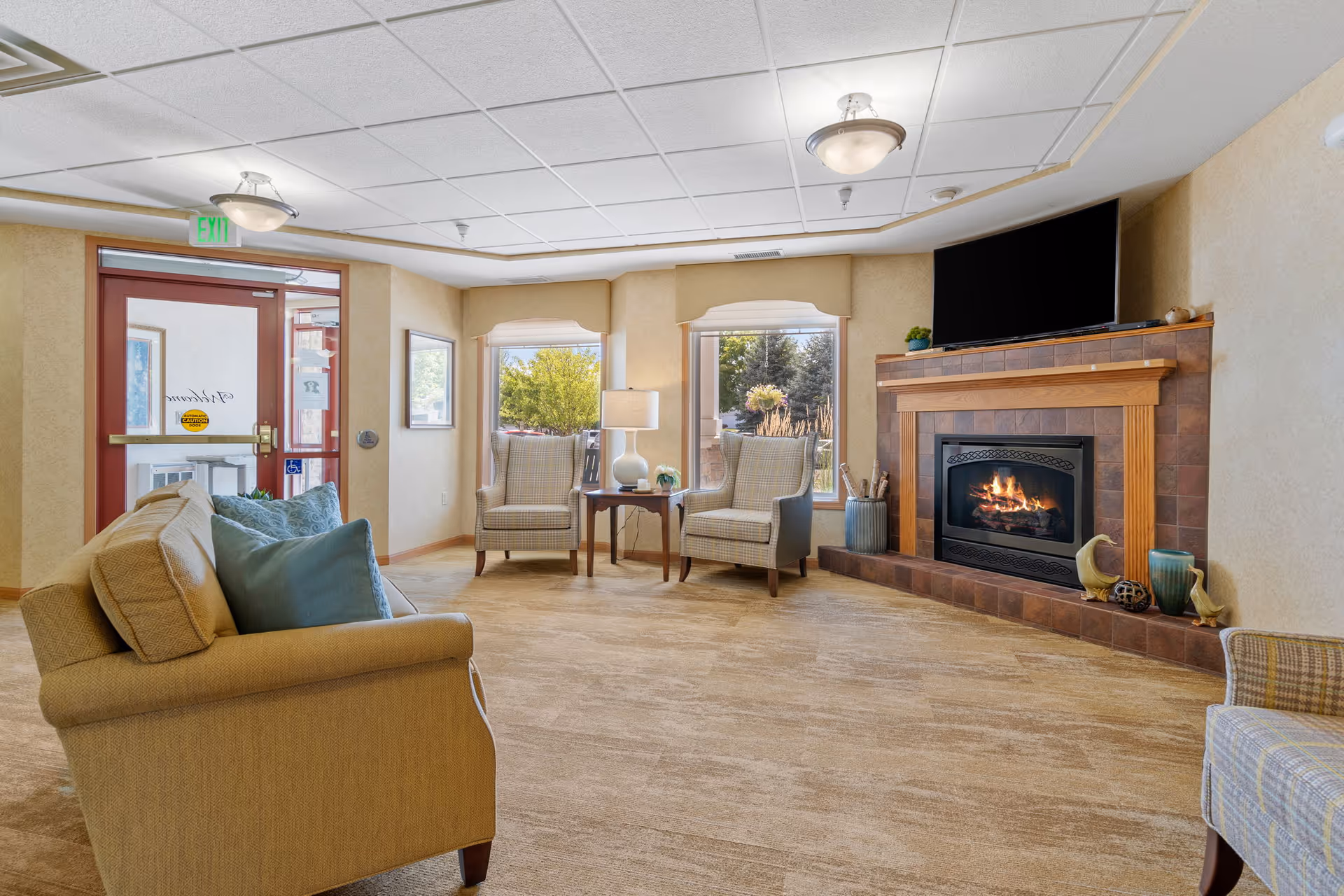 Sunny lounge with sofas and armchairs arranged around a lit fireplace and wall-mounted TV near an entry door.