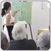 A woman standing and painting on a canvas mounted on an easel while several elderly people watch her from their seats.