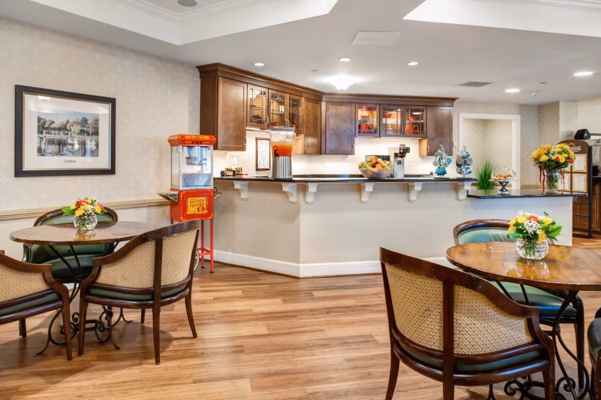 Bright communal dining area with round wooden tables and chairs in front of a service counter and kitchenette with cabinets and a popcorn machine.