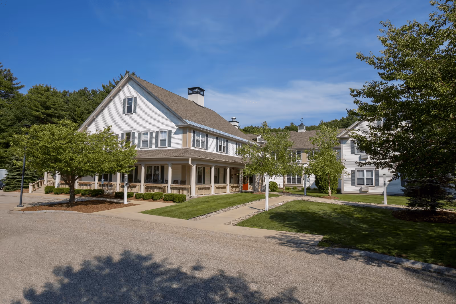 Exterior view of a large, white, multi-story senior living facility building with a wrap-around porch, surrounded by green trees and well-maintained lawns under a clear blue sky.