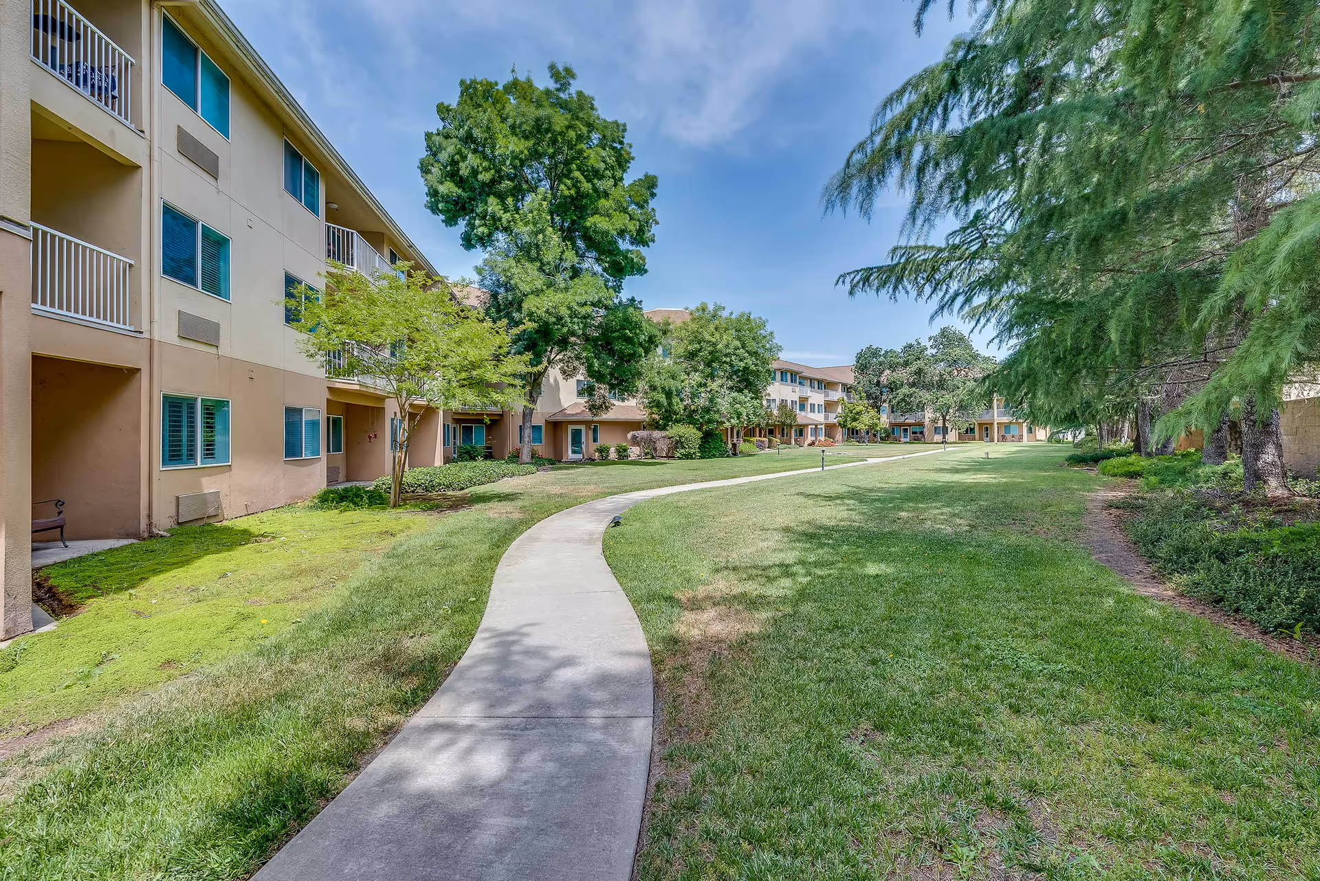 A winding concrete pathway runs through a grassy courtyard area surrounded by a multi-story senior living facility building. The building has beige and light brown walls with blue window shutters and balconies. There are several trees and shrubs providing greenery and shade under a partly cloudy blue sky.