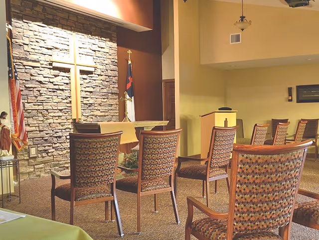 Interior of a small chapel or worship room with a stone wall featuring a large wooden cross, an altar with a cloth, several patterned chairs arranged facing the altar, and flags on either side of the altar.