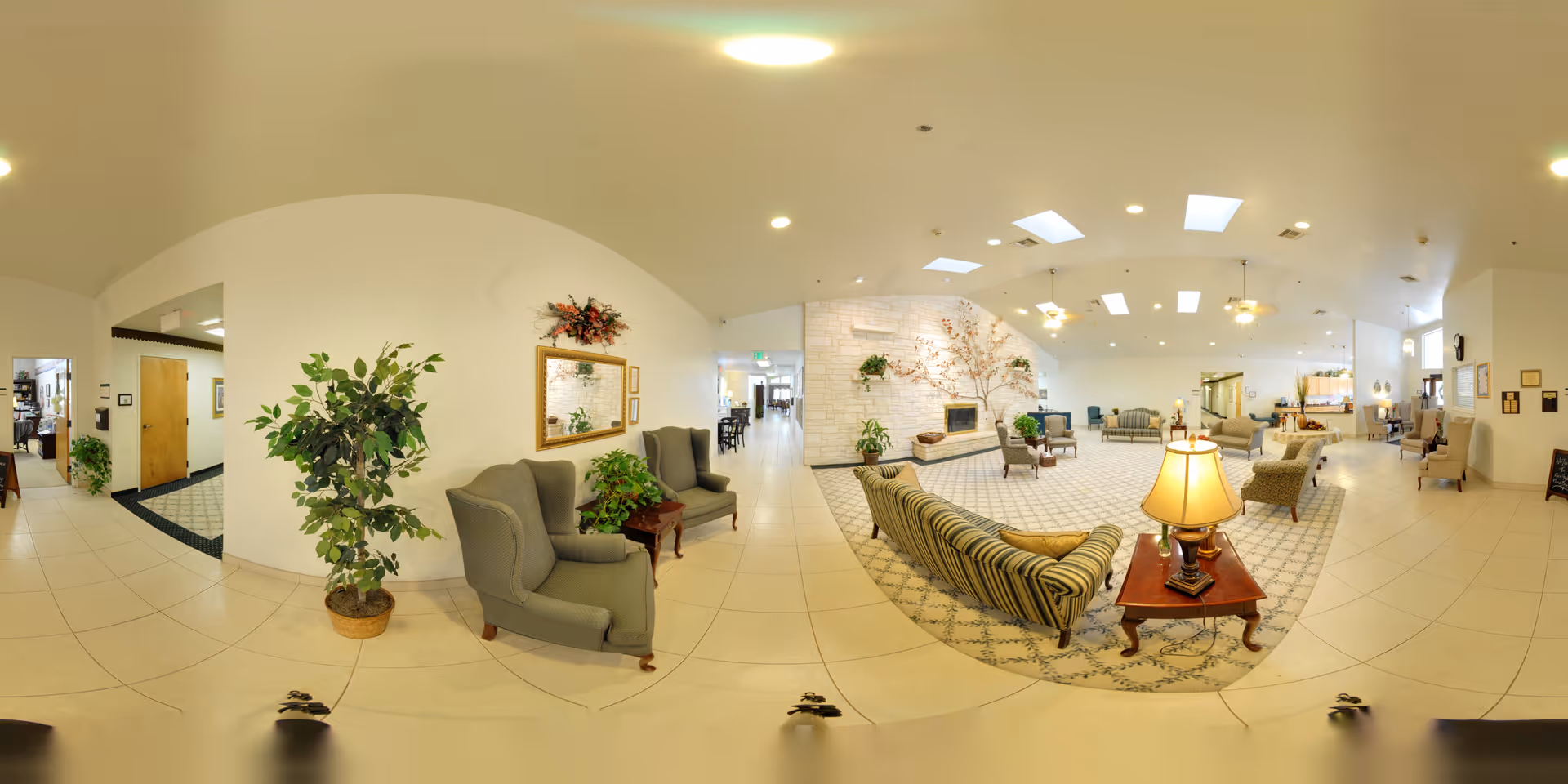 A spacious and well-lit senior living facility common area with comfortable seating including armchairs and sofas arranged around a central coffee table with a lamp. The room features a large stone fireplace decorated with plants and floral arrangements. The ceiling has multiple skylights and recessed lighting, and the floor is tiled with a patterned carpet in the seating area. There are plants and framed mirrors on the walls, and a hallway leading to other rooms is visible in the background.