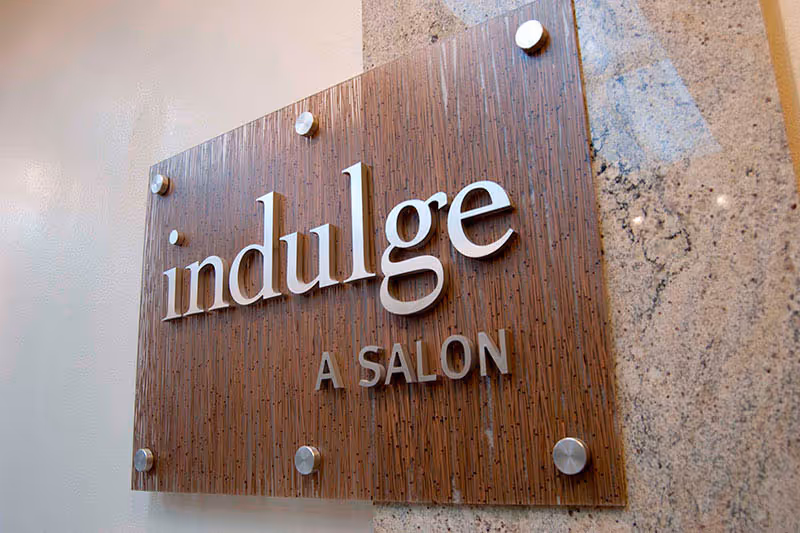 A wooden and glass wall sign with the words 'indulge A SALON' mounted on a wall with a granite surface on the right side.